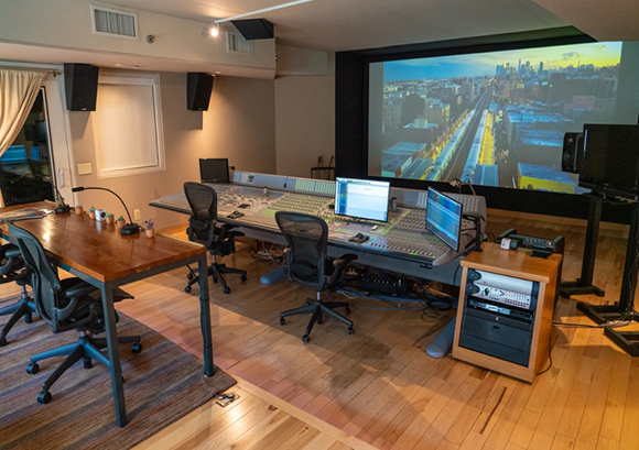 A professional audio mixing studio featuring a large console, computer monitors, and a large projection screen.