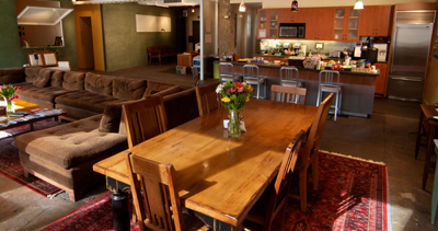 A large wooden dining table with chairs sits in front of a brown sectional sofa and a kitchen area.