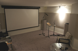 A large white projector screen, a microphone on a stand, and a speaker in a dimly lit room.