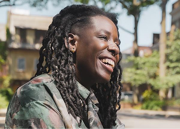 A smiling person with long dark dreadlocks wearing a camouflage shirt outdoors.