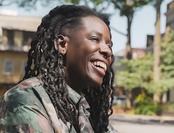 A smiling person with long dark dreadlocks looking off to the side outdoors.