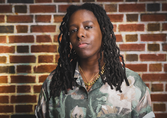 A person with long dark dreadlocks and a nose ring wearing a patterned shirt against a brick wall.