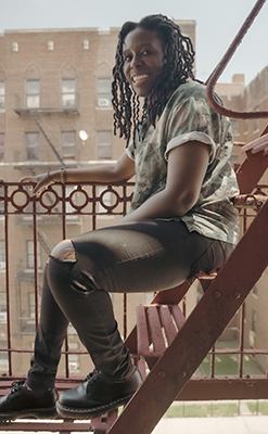 A smiling woman with long braids sits on red metal fire escape stairs in front of a brick building.