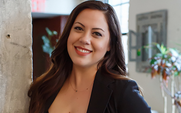 A smiling woman with long brown hair wearing a black blazer in an indoor setting.