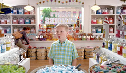 A young boy in a plaid shirt stands in a brightly lit candy shop filled with jars of sweets.