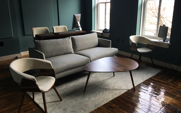 A grey sofa and wooden coffee table sit on a light rug in a room with teal walls and large windows.
