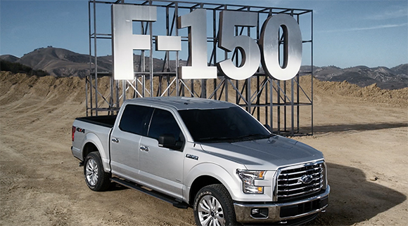 A silver Ford F-150 pickup truck parked in a dirt lot before a massive metal F-150 sign.