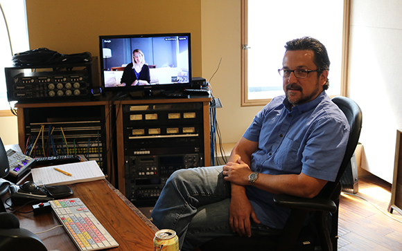 A man in a blue shirt sits in an office with a monitor showing a woman on a video call.
