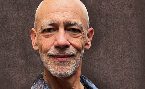 Close-up portrait of a bald man with grey facial hair and a slight smile against a textured brown background.