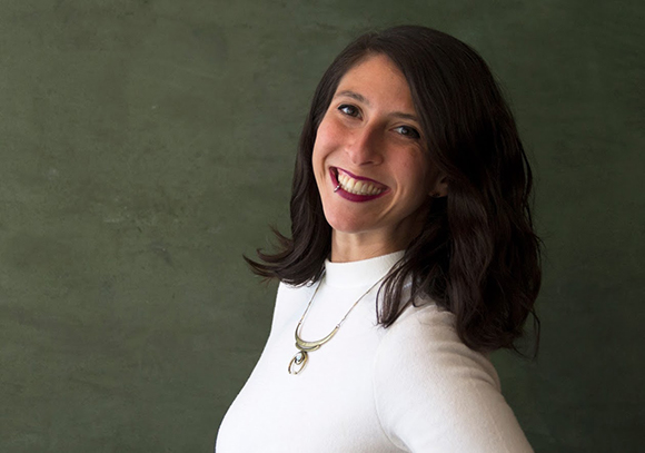 A smiling woman with dark wavy hair wearing a white turtleneck and a silver necklace.