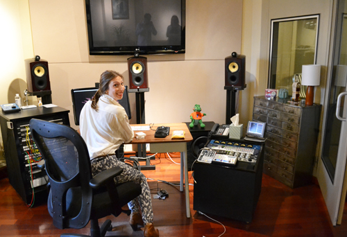 A woman sits in an office chair at a desk surrounded by speakers and audio mixing equipment.