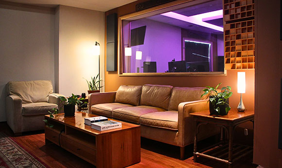A living room with a leather sofa and coffee table looking through a window into a purple-lit studio.