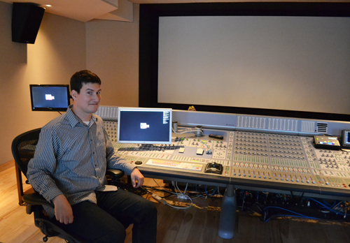A man sits in a chair behind a large professional audio mixing console in a studio with a projection screen.