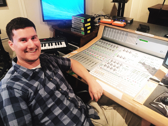 A man in a plaid shirt sits at a large audio mixing desk in a studio with a monitor and keyboard.