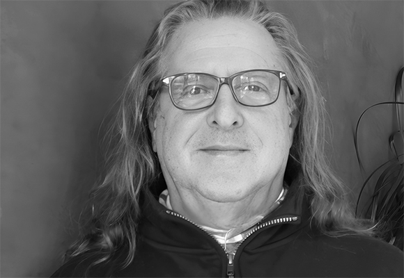 Black and white close-up portrait of a man with long hair wearing glasses and a dark zip-up jacket.