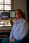 A man with long hair and glasses sitting in a room with multiple computer monitors and studio equipment.