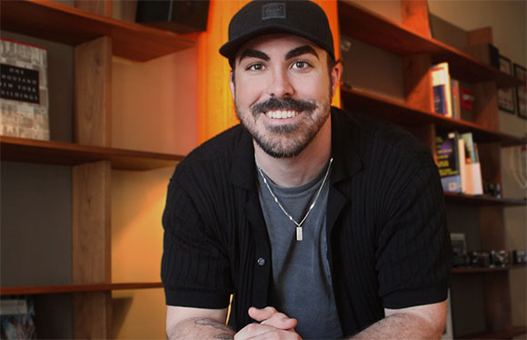 A smiling man with a beard and black baseball cap wearing a black shirt over a grey t-shirt.
