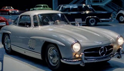 A vintage white Mercedes-Benz coupe parked in a museum showroom with other classic cars in the background.