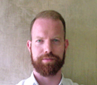 A man with short light hair and a full reddish-brown beard wearing a white shirt.