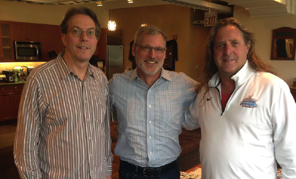 Three men smiling and standing side-by-side in a kitchen or living area.