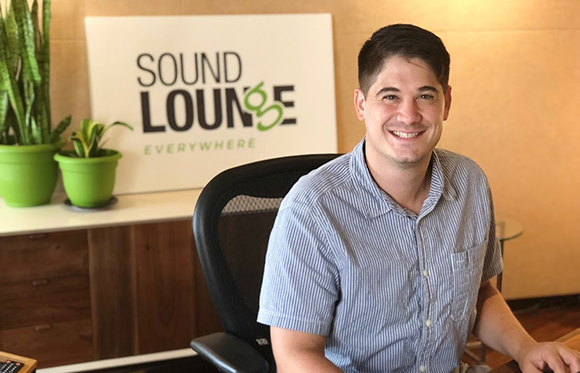 A smiling man in a blue striped shirt sitting in an office chair in front of a Sound Lounge sign.