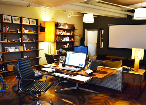 A workspace featuring a wooden desk, computer monitor, black leather chair, and various lamps.