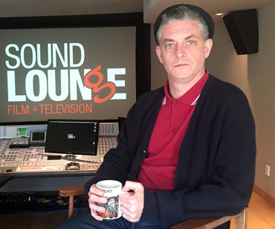 A man in a red polo and black cardigan sits in a studio holding a mug in front of a Sound Lounge screen.