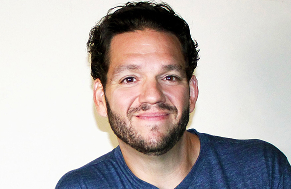 A man with dark wavy hair and a beard smiling against a plain white background.