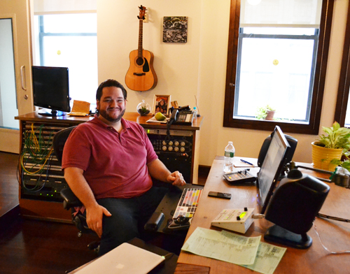 A smiling man in a red polo shirt sits in an office filled with computers, audio gear, and a guitar on the wall.