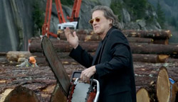 A man in a black suit and sunglasses holds a large chainsaw amidst piles of logs.