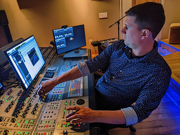 A man in a patterned shirt adjusts knobs on a large professional audio mixing console with multiple monitors.
