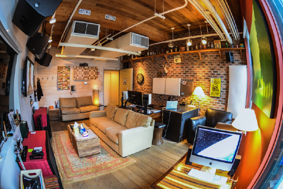 A wide-angle shot of a living room with brick walls, wooden ceilings, leather sofas, and a computer desk.