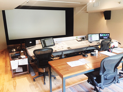 A large mixing console sits before a massive projection screen in a professional recording studio.