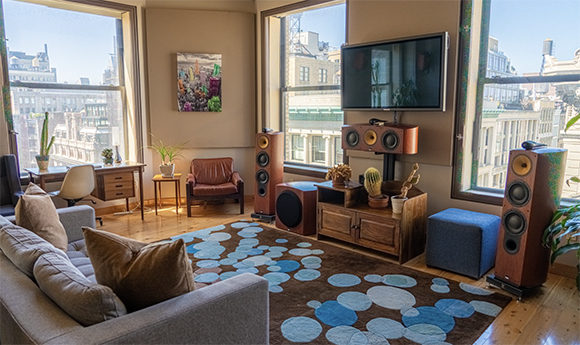 Living room with large windows, a grey sofa, a patterned rug, and large wooden floor-standing speakers.
