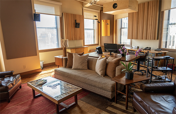 A living room featuring a brown sofa, wooden coffee table, leather armchairs, and wall-mounted speakers.