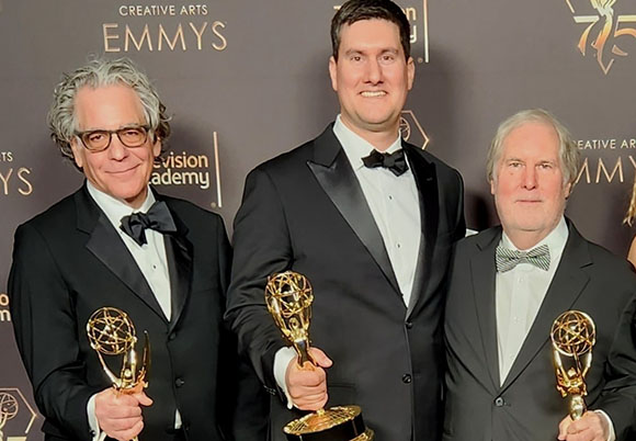 Three men in formal black tuxedos posing together while holding gold Emmy awards.