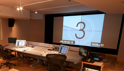 A broadcast control room with a large projection screen showing a countdown and multiple mixing consoles.