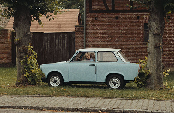 A light blue vintage microcar parked on grass between two large trees in front of a brick building.