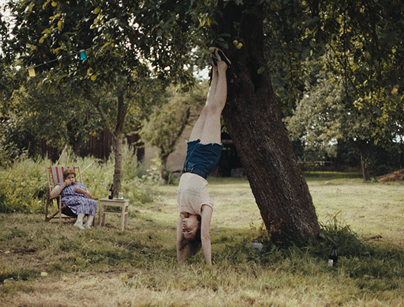 A child does a handstand against a large tree trunk in a grassy yard while a woman sits in a chair nearby.