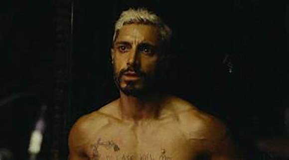 A shirtless man with short bleached hair and a dark beard looking intensely into the distance.