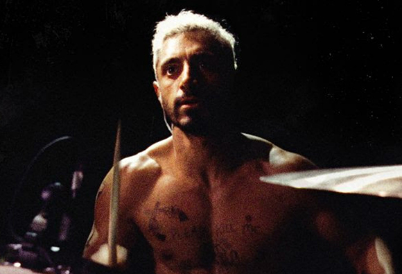 A shirtless man with light hair plays drums in a dark setting under a spotlight.