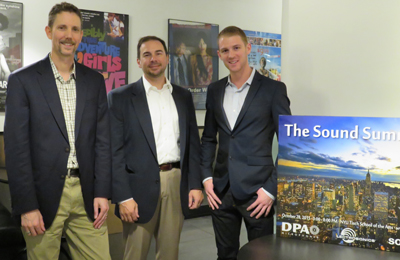 Three men in business casual attire standing in an office with posters in the background.