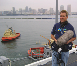 A man on a boat holds a large, fuzzy cylindrical device with a city skyline and tugboat in the background.