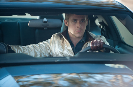 A man with short blonde hair wearing a gold jacket sits behind the steering wheel of a car.