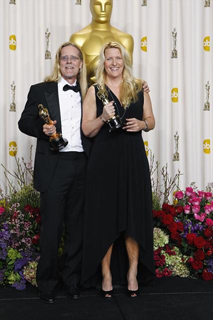 A man in a tuxedo and a woman in a black dress hold gold awards in front of an Oscar backdrop.