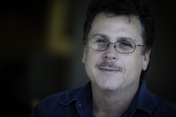 A man with dark wavy hair, glasses, and a mustache smiling slightly against a dark background.
