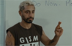 A man with bleached hair and a beard wearing a brown tank top stands before a whiteboard.