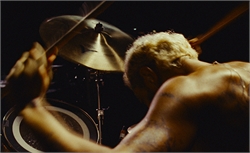 A shirtless person with blonde hair playing a drum set with sticks in motion.
