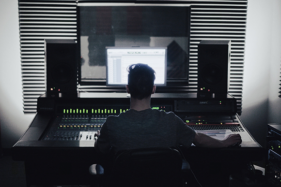 A person sits at a large mixing console in a dark studio with monitors and computer screens.