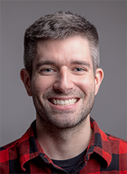 A smiling man with short salt-and-pepper hair and stubble wearing a red and black plaid shirt.